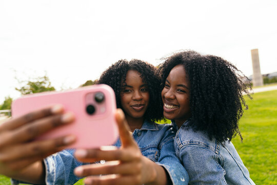 Two cheerful twin sisters are taking a selfie with a smartphone in a park, enjoying their time together