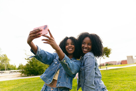 Two happy twin sisters are taking a selfie with a smartphone in a park, enjoying their time together