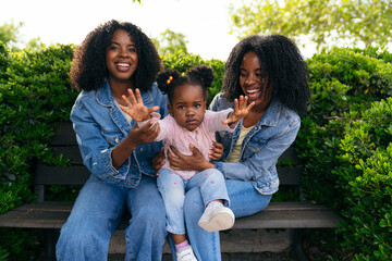 Twin sisters smiling and holding their niece, enjoying a sunny day in the park while creating...