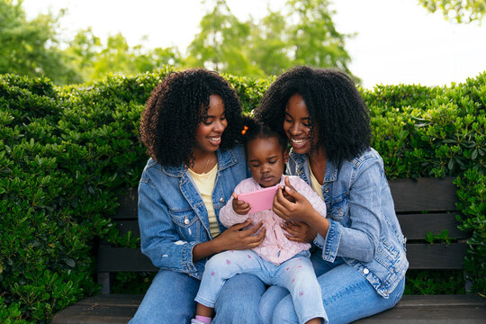 Two twin sisters enjoying time outdoors with their niece, sharing a smartphone on a park bench, creating a heartwarming family moment