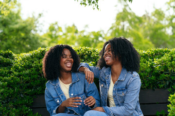 African two happy twin sisters are sitting on a park bench, wearing denim jackets and laughing together on a sunny day
