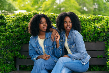 Two twin sisters wearing denim jackets are holding hands while sitting together on a park bench...