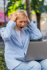 Upset mature businesswoman reading bad news on laptop sitting on bench on city street. Frustrated...