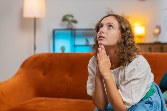 Sad young woman sitting on sofa at home in shock by awful news, folding palms together pray ask for help, thinks feel stressed looks desperate by life problems. Praying, asking for help. Lifestyles.