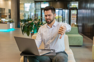 Dislike. Upset businessman guy working on laptop computer in office lobby showing thumb down sign gesture, expressing discontent, disapproval, dissatisfied bad work. Displeased serious man in hotel.
