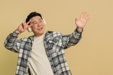 Happy Asian middle-aged man listening music on headphones and dancing disco fooling around having fun expressive gesticulating hands celebrating. Mature adult guy isolated on beige background indoors