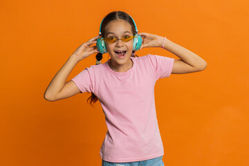 Happy smiling preteen child girl in headphones listening music dancing disco fooling around having fun expressive gesticulating hands relaxing on party making funny moves. Kid on orange background