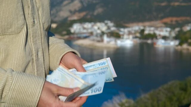 Man counting turkish lira banknotes by the sea. Tourist counting turkish lira banknotes while standing near coastal scenery with seascape and seaside town buildings under blue sky