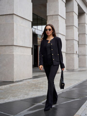 Elegant woman in sleek black outfit, consisting of a jacket and pants, walks confidently on stone pavement. She carries a stylish handbag, wears dark sunglasses, and her hair is loose