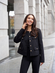 Smiling woman with long dark hair touches her face while wearing a stylish black outfit, jacket...