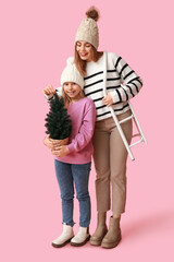 Cute little girl with small Christmas tree and her mother holding sled on pink background