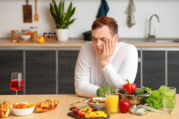 Young sad overweight man choosing healthy food over fast food at table in kitchen. Weight gain...