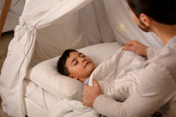 Father putting his little son to bed in hut at home