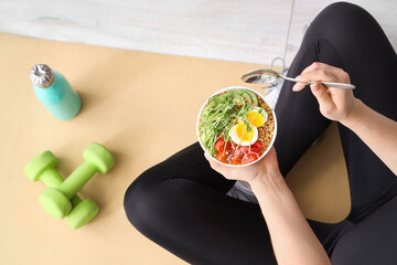 Sporty young woman sitting on yoga mat with bowl of nutritious oatmeal and dumbbells at home, top...