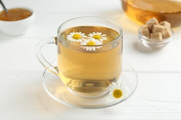 Delicious chamomile tea in glass cup, fresh flowers, brown sugar and jam on white wooden table,...