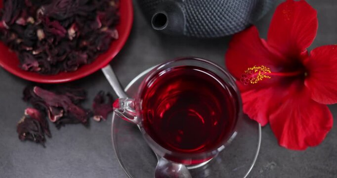 Tasty hibiscus tea in cup, teapot, dry roselle sepals and beautiful flower on grey table, camera moving