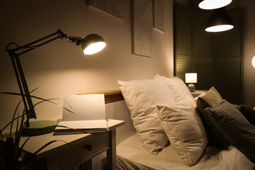 Glowing lamp with book on table in bedroom at night