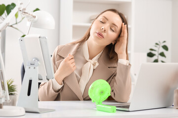 Young businesswoman with mini fan suffering from heat at table in office