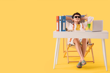 Young businessman waiting for summer vacation in deck chair at table on yellow background