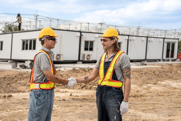 Two construction workers wearing hard hats and safety vests shaking hands at a construction site,...
