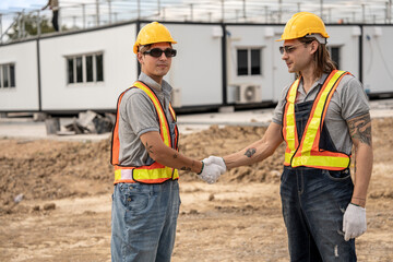 Two construction workers wearing hard hats and safety vests shaking hands at a construction site, symbolizing collaboration and successful teamwork with modular units in the background.