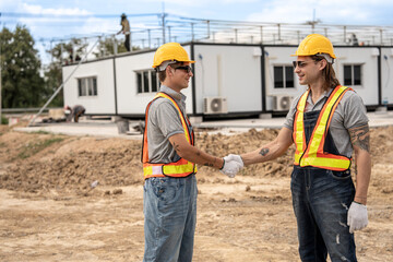 Two construction workers wearing hard hats and safety vests shaking hands at a construction site,...
