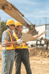 Workers in safety vests and helmets reviewing construction plans at a job site, highlighting...