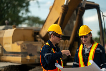 Two construction workers in hard hats and reflective vests discussing blueprints at an industrial...