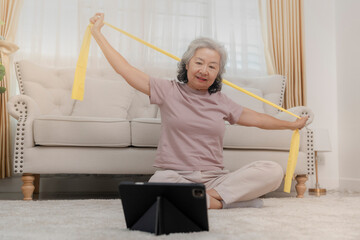An elderly woman exercises with a resistance band at home, focusing on health and fitness. She...