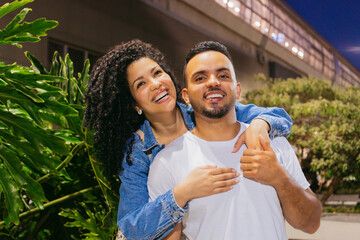 Young Latin man smiles as his cheerful girlfriend hugs him from behind in a flowery cityscape