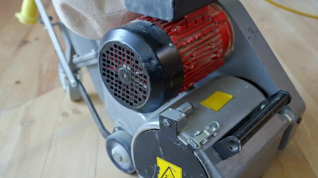 Electric wood floor sanding machine with red and silver motor, dust bag attached, camera pans over the equipment on pine parquet floor during home renovation, highlighting tool components,