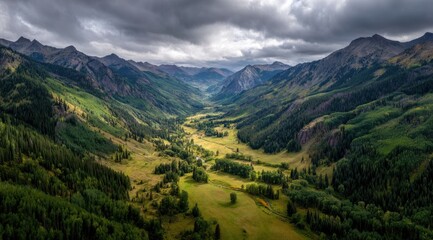Obraz premium An aerial view of a verdant valley nestled between majestic mountains, under a dramatic, partly cloudy sky. The valley floor shows a mix of meadow and forest, with a river snaking through