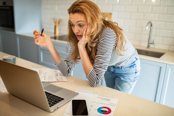 Woman working remotely from kitchen while drinking coffee and reviewing documents on laptop