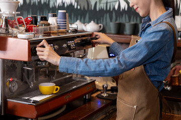Asian female barista in brown apron and denim shirt operating espresso machine with focused expression. Yellow coffee cup placed under portafilter as steam rises. Professional café preparation moment.