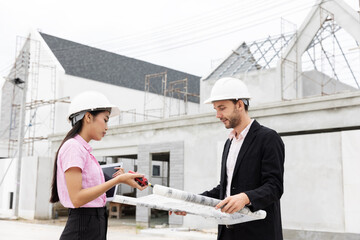 Two architects wearing safety helmets discuss blueprints at a construction site. Surrounded by scaffolding and a partially built structure, they collaborate on the details of the building project.