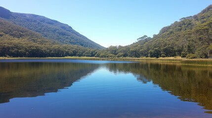 Fototapeta premium Serene mountain lake, reflecting a blue sky. Lush green hills flank the tranquil water