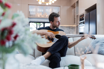 A man relaxes at home on a cozy couch while playing an acoustic guitar, surrounded by a modern kitchen and warm lighting. The atmosphere is calm and inviting, perfect for a quiet music session.
