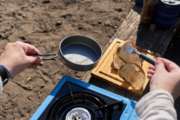 Unrecognizable person toasts bread outdoors, using a frying pan on a gas portable stove, for breakfast at his campsite.
