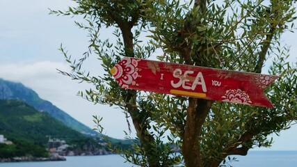 Wooden road sign on green olive tree with saying I SEA YOU joke pun. Concept of summer holiday vacation on seacoast. Idea of tropical travel tourism taking a break wellbeing slowing life. 