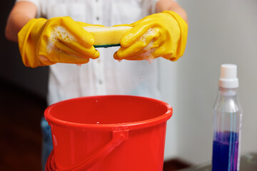 Woman in yellow gloves holding sponge over red bucket and cleaning solution bottle