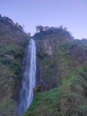 Tall waterfall cascading down lush green cliffs under sunlight, creating a serene and majestic view.