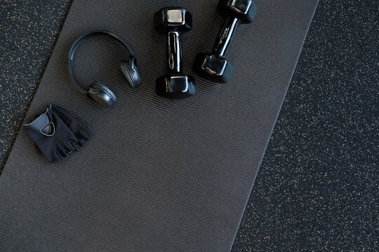 Headphones and black dumbbells lie on a black yoga mat. Flat lay. Space for text.