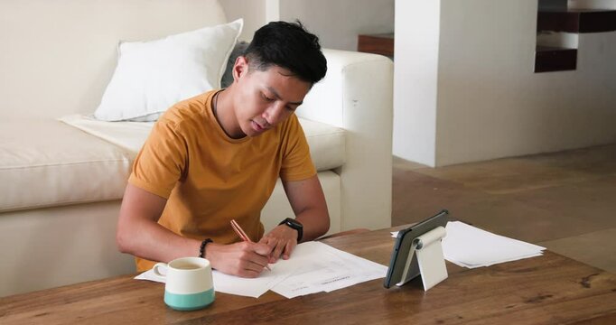 Young man writing on paper at home, using tablet for reference