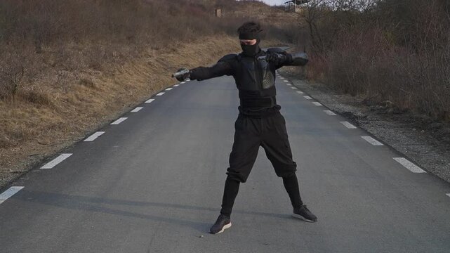 A figure in a black ninja costume strikes a pose with guns on a deserted asphalt road under a clear sky. Dramatic and action-oriented.