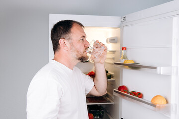 Man enjoying refreshing water from a glass while standing near an open refrigerator filled with...