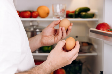 Hands holding fresh eggs in a refrigerator filled with colorful vegetables fruits, showcasing the...