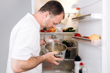 Man inspecting a pot of spoiled food in an open refrigerator filled with various vegetables and...