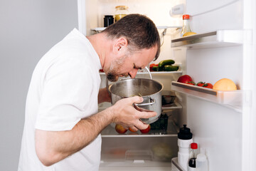 Man inspecting a pot of spoiled food in an open refrigerator, surrounded by various fruits and...