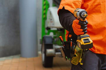 Close-up of a construction worker with a tool belt, wearing protective gloves, holding a cordless drill. Concept on a construction site with green industrial equipment.