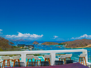 Fototapeta premium An outdoor dining area with a wooden table and chairs, overlooking the sea with mountains in the distance, under a clear blue sky. This serene setting is perfect for enjoying Labuan Bajo’s beauty